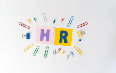 Letters ‘H’ and ‘R’ on coloured cards, surrounded by colourful paper clips, drawing pins and staples on a white background.