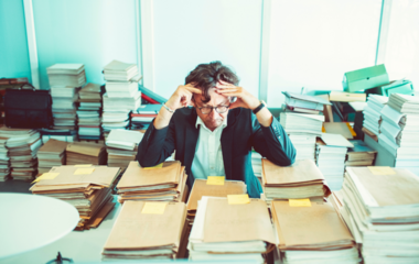  A stressed businessman sits in an office surrounded by piles of files and documents.