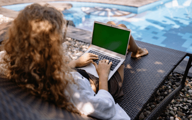 A woman sits on a lounger by the pool and works on her laptop. 