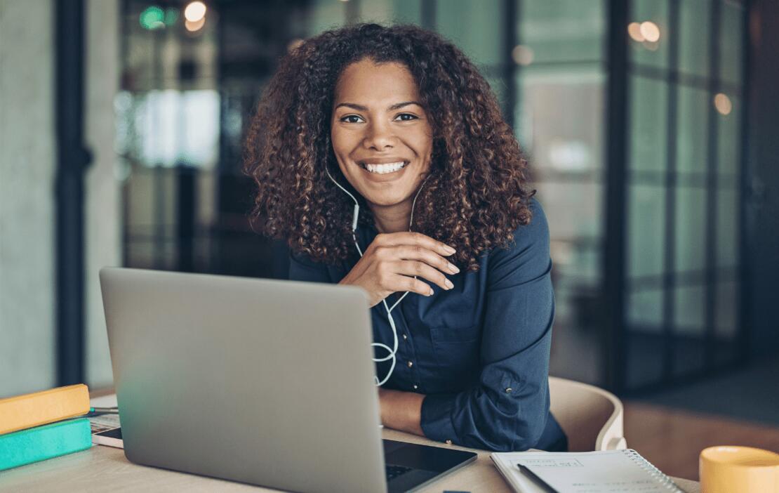 A woman with curly hair sits in front of her laptop and smiles 