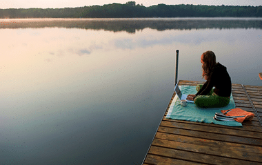 Eine Frau sitzt auf einem Steg am Wasser auf einer Decke und arbeitet 